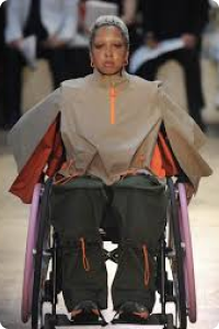 Woman in a wheelchair wears beige coat with orange lining and zip at neck on a catwalk